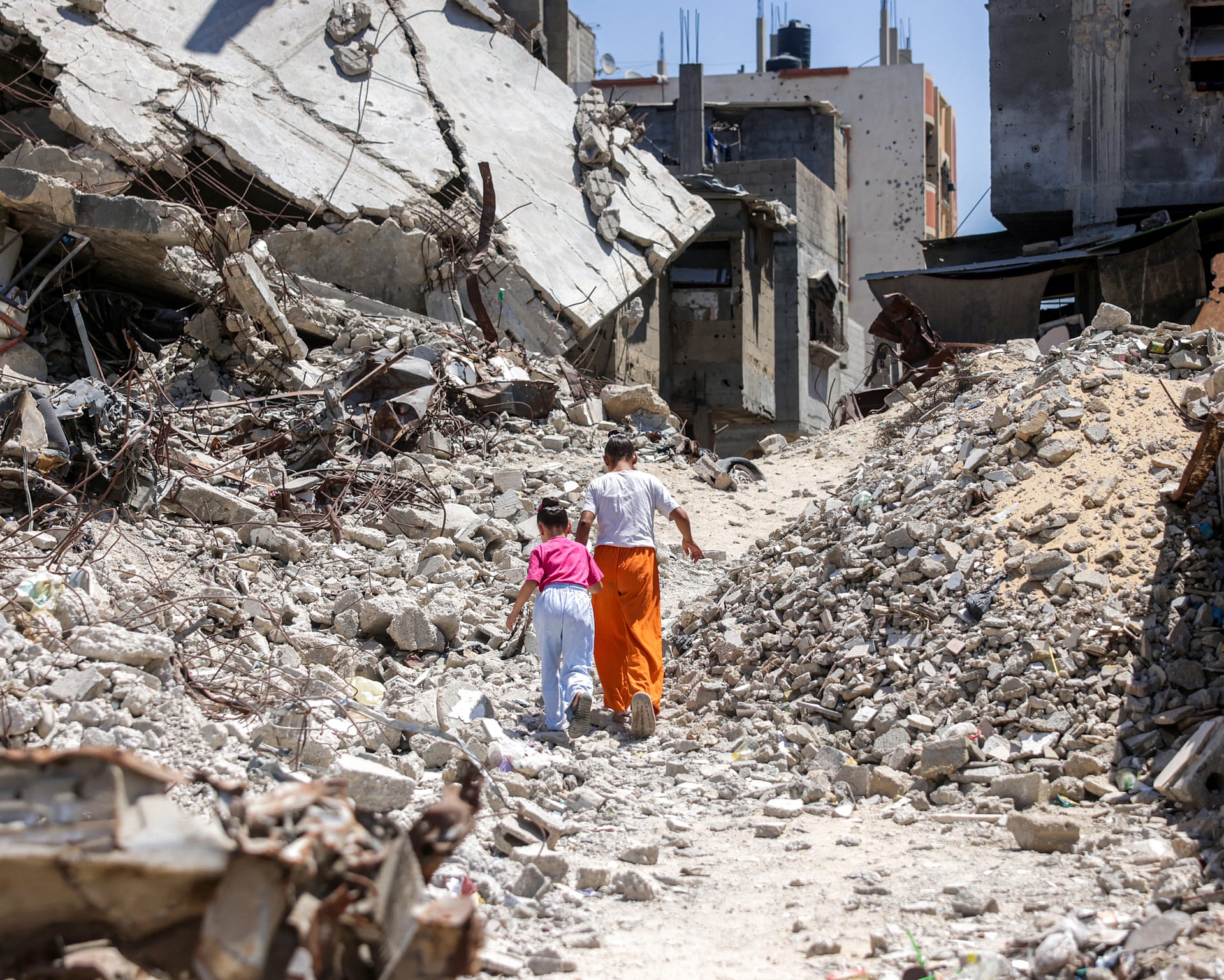 Two girls walk through a landscape of collapsed concrete and rubble in Gaza under a blue sky.