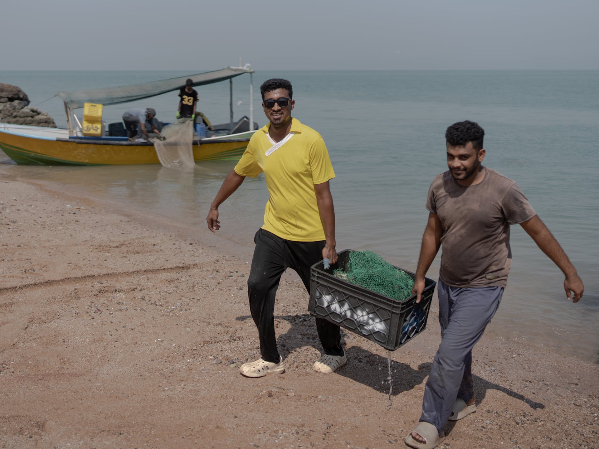 Two fishermen carry a crate of catch along a beach near Bandar Abbas, with a small boat behind them.