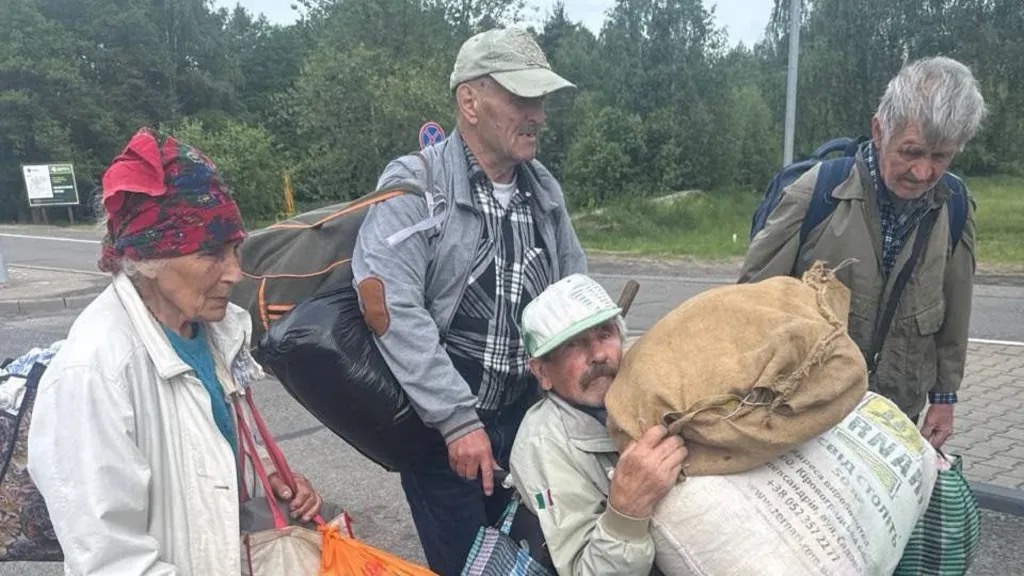 Four elderly residents carrying sacks and bags after leaving Oleshky, standing beside a road.
